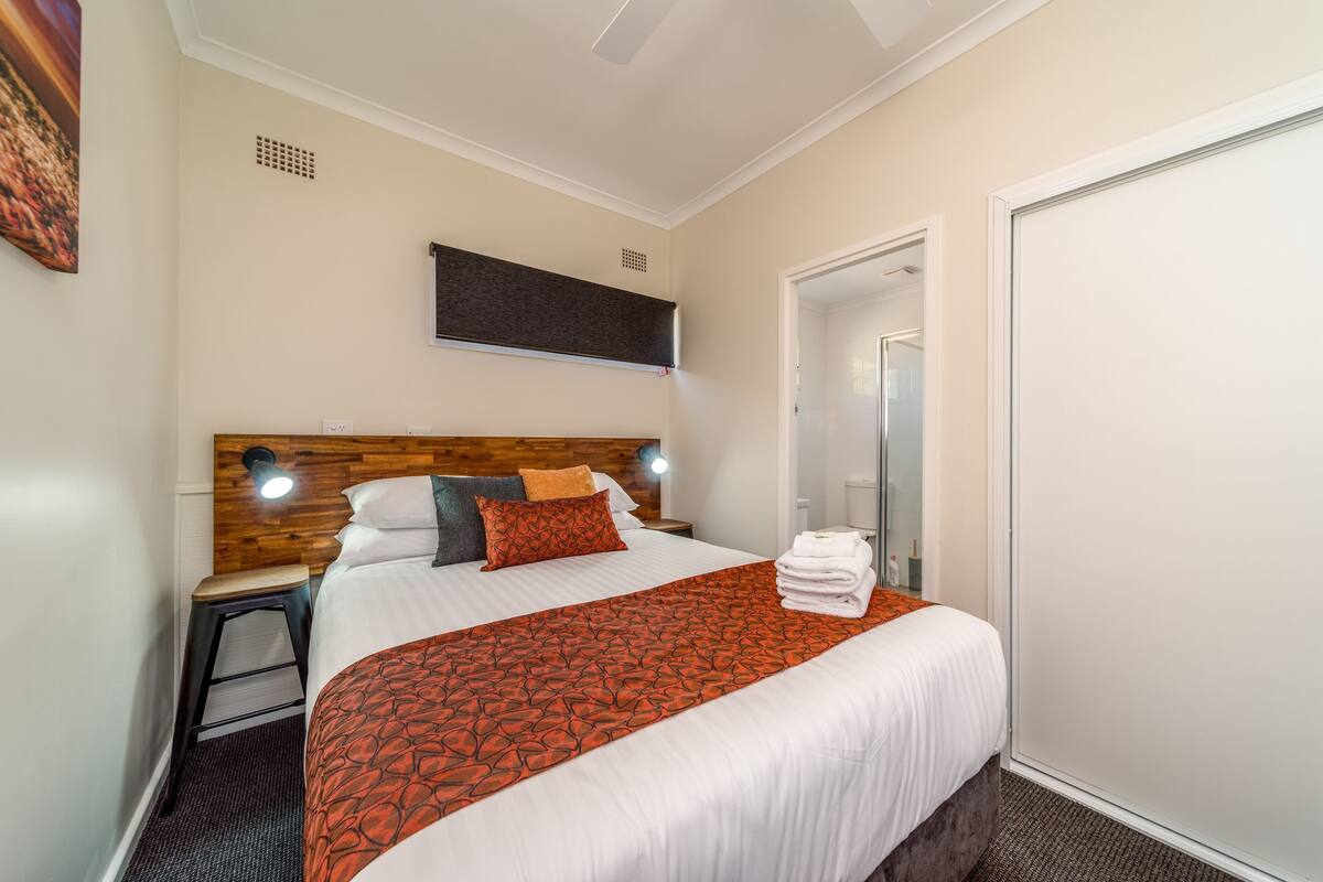 Een keurig opgemaakt bed met een bedspreider met een patroon, een houten hoofdbord en een nachtkastje met een lamp.