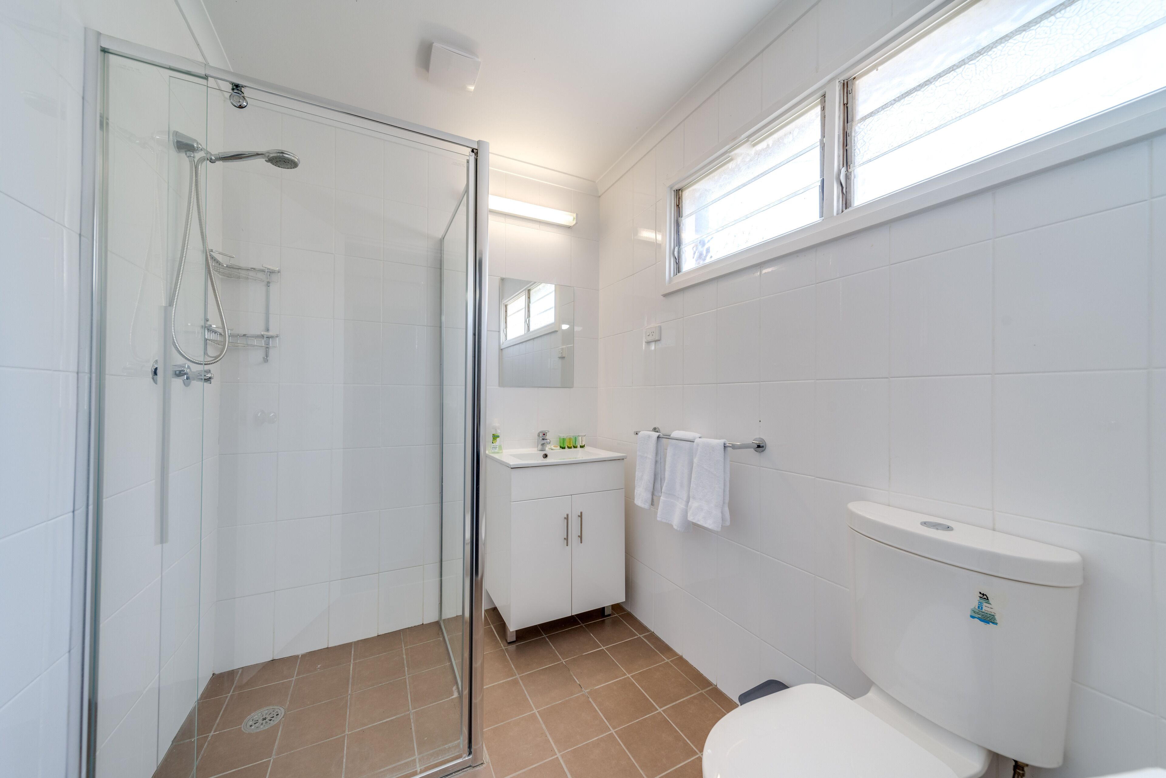 A modern bathroom with a glass shower enclosure, a toilet, a sink with a mirror above, and a window providing natural light.