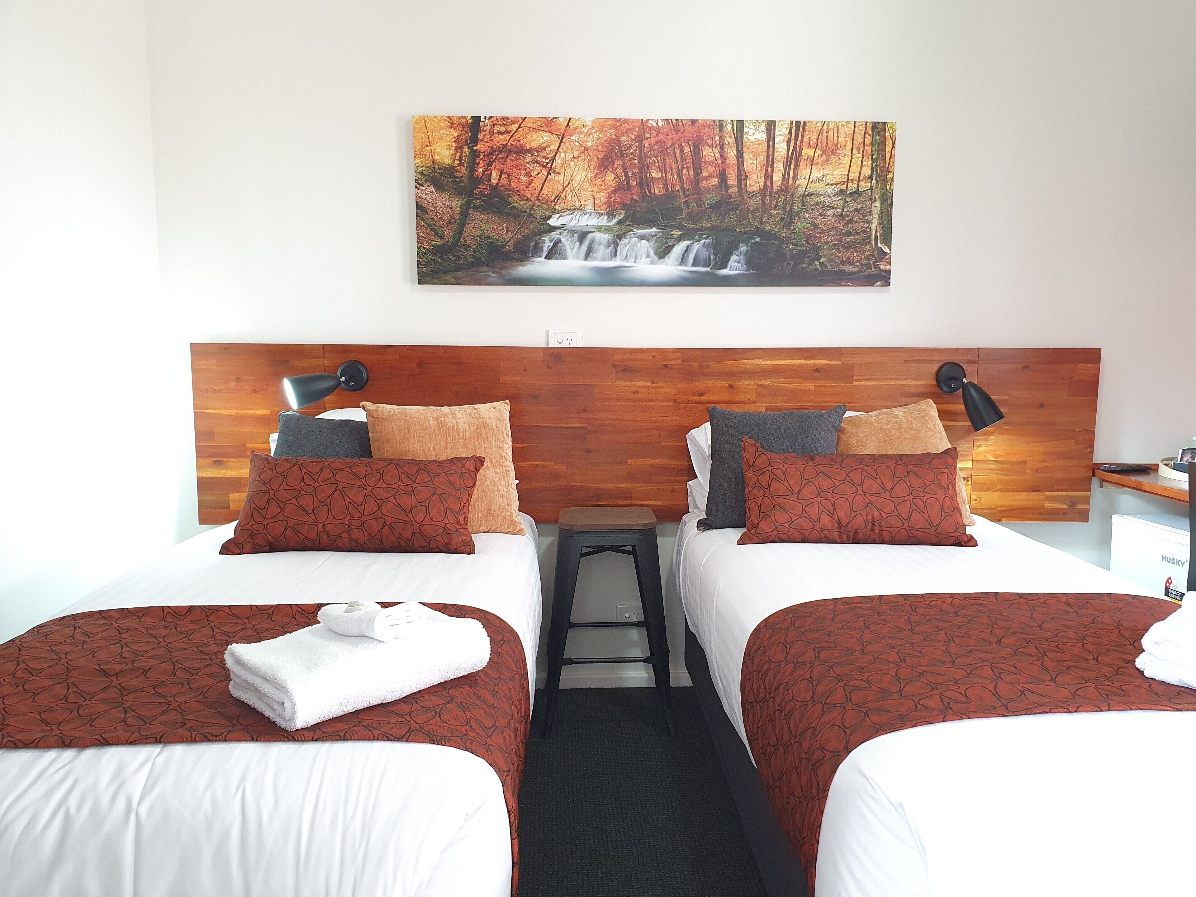 Habitación de hotel con dos camas, cabecera de madera, un cuadro de una cascada y un escritorio pequeño con una lámpara.