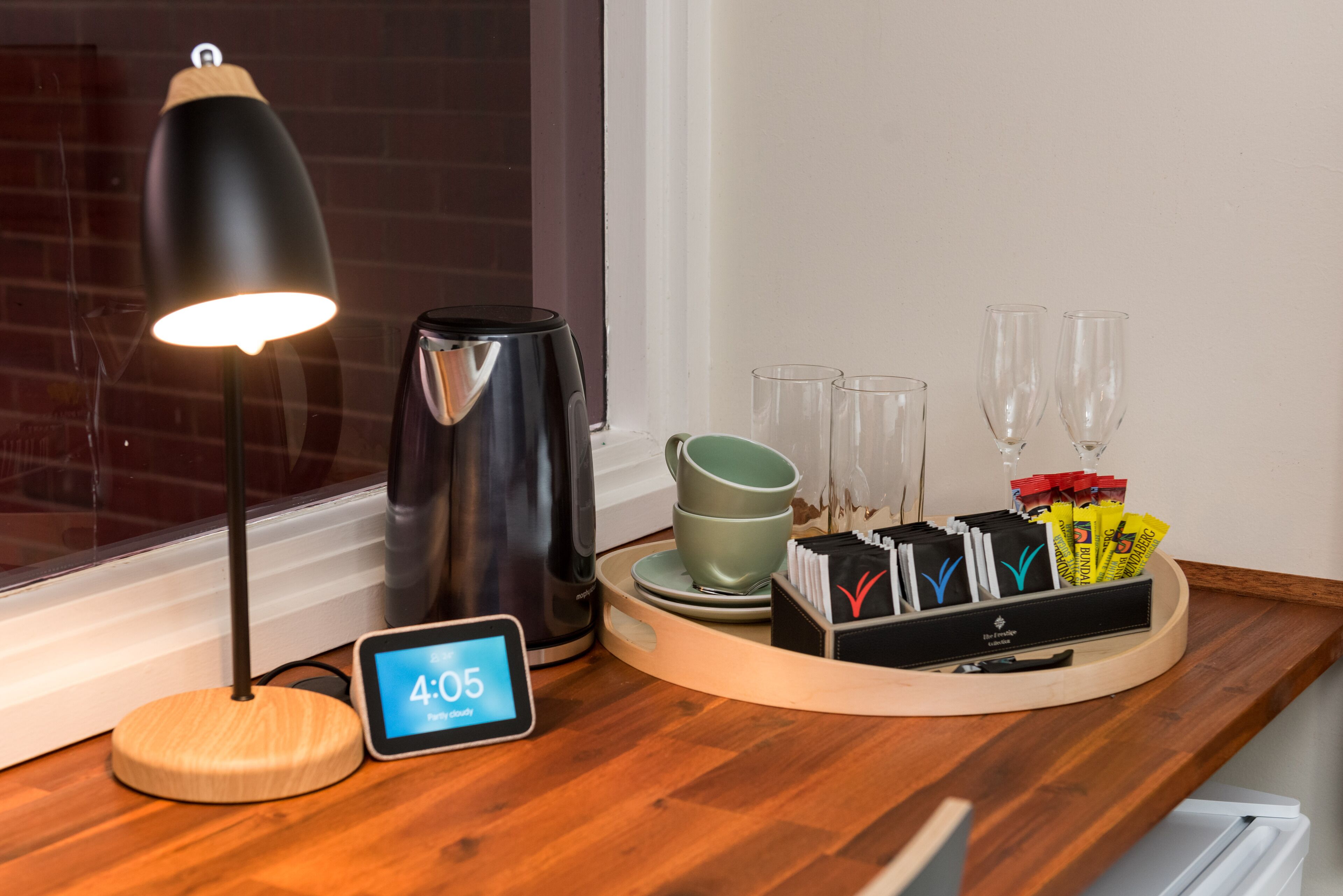 Een houten tafel met een koffiezetapparaat, een klok waarop 4:05 staat, een lamp en een dienblad met kopjes en servetten.