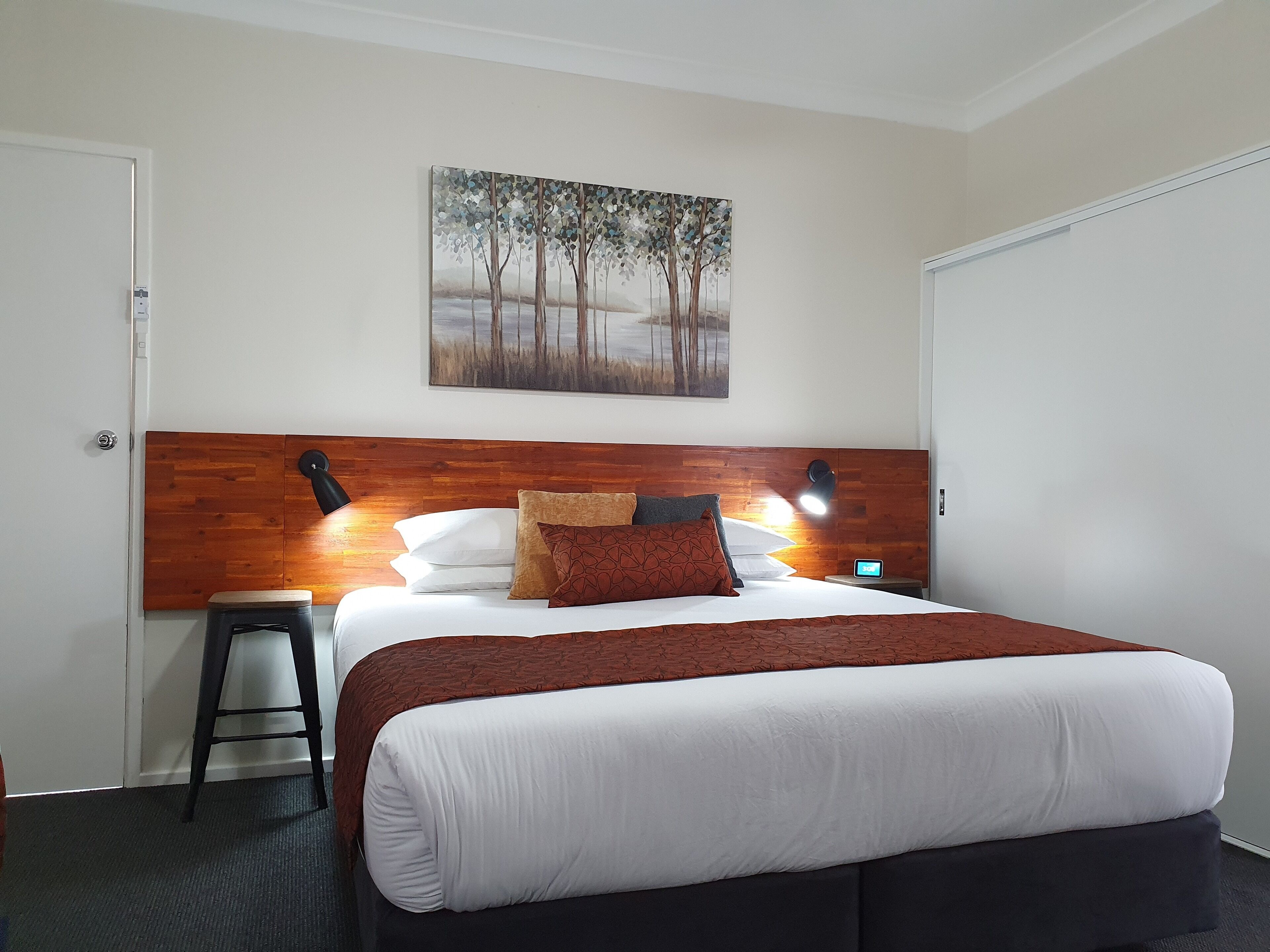 Ein Hotelzimmer mit einem hölzernen Kopfteil, einem Bett mit weißer Bettwäsche und einer braunen Decke, einem Nachttisch mit Lampe sowie einem Baumgemälde an der Wand.