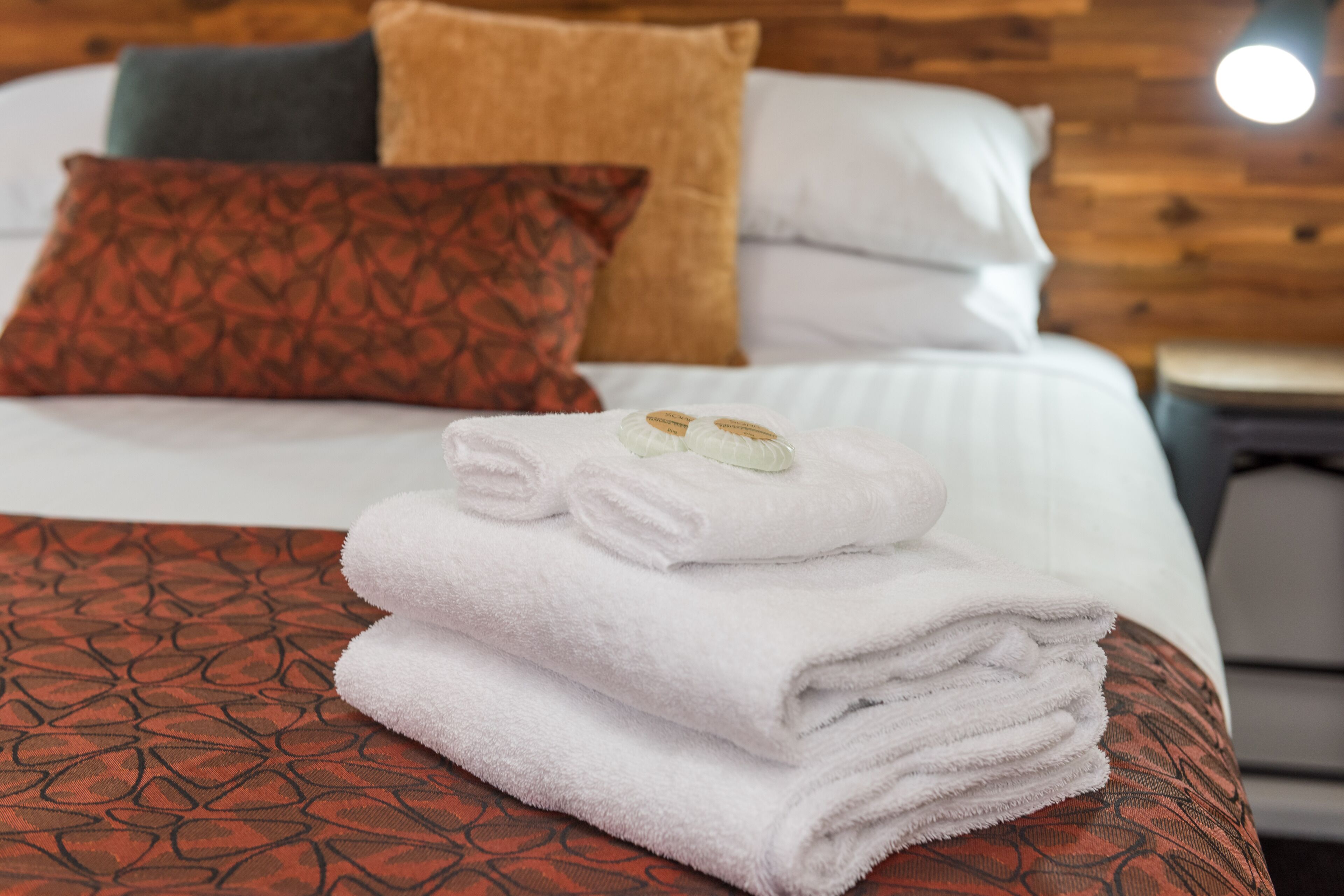 Un lit bien fait, avec une literie blanche et à motifs, une pile de serviettes blanches pliées et un coussin décoratif à motifs rouges.