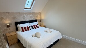 Een slaapkamer met dakraam, een bed met witte beddengoed en rode kussens, een nachtkastje met lamp, en een vloerbedekking.