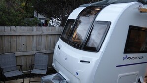 Een witte camper met de merknaam 'BAILEY' en 'PHOENIX' zichtbaar, geparkeerd naast een houten hek en afgedekt met een beschermhoes.
