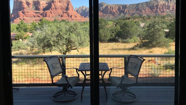 A balcony with two chairs and a table, overlooking a mountainous landscape with red rock formations and green trees.