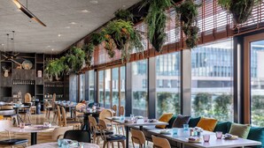 Een modern restaurant met hoge plafonds, hangende planten en grote ramen met uitzicht op een stadsbeeld.