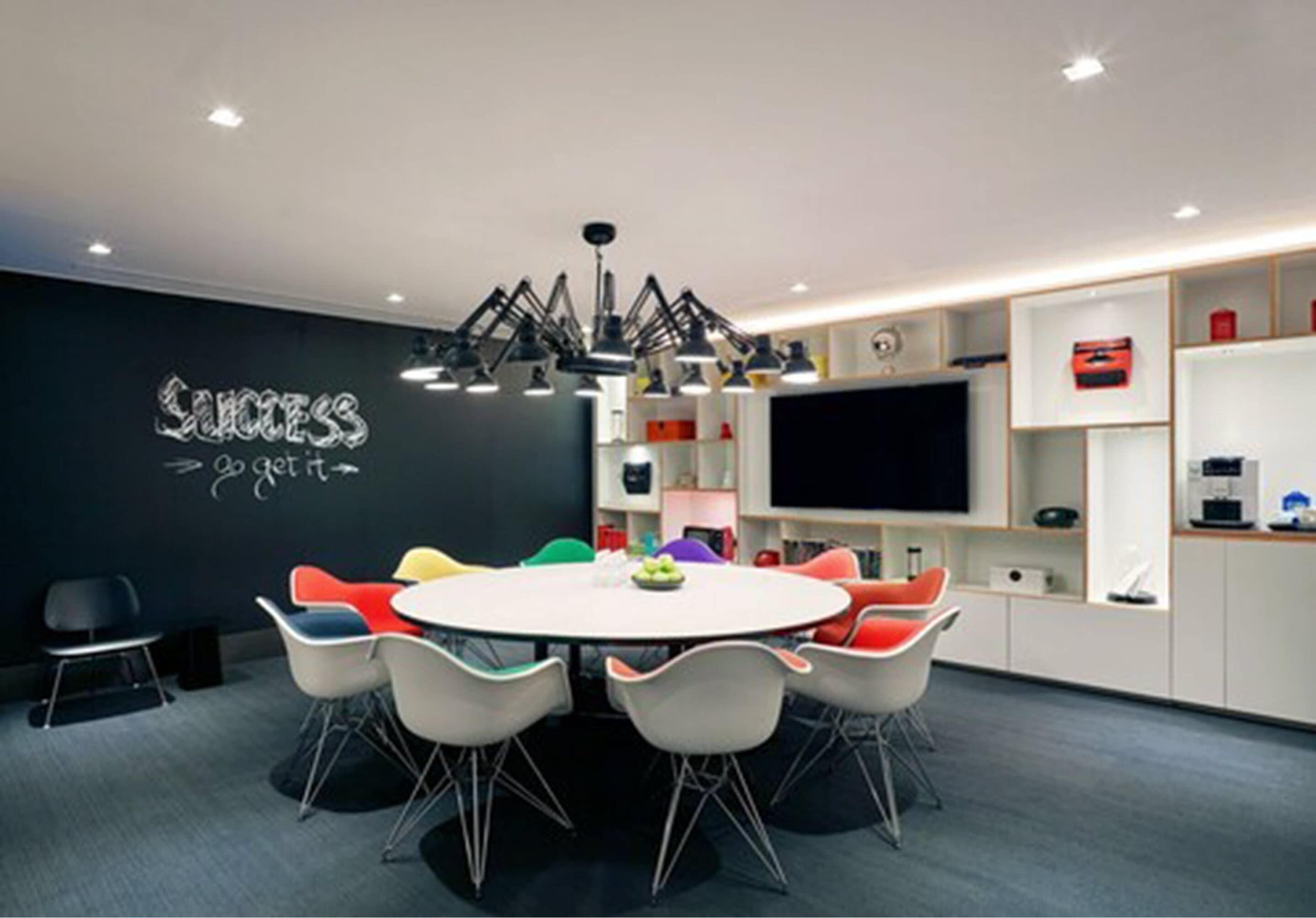 A modern meeting room with a round table, colorful chairs, a blackboard with motivational writing, and a flat-screen TV.