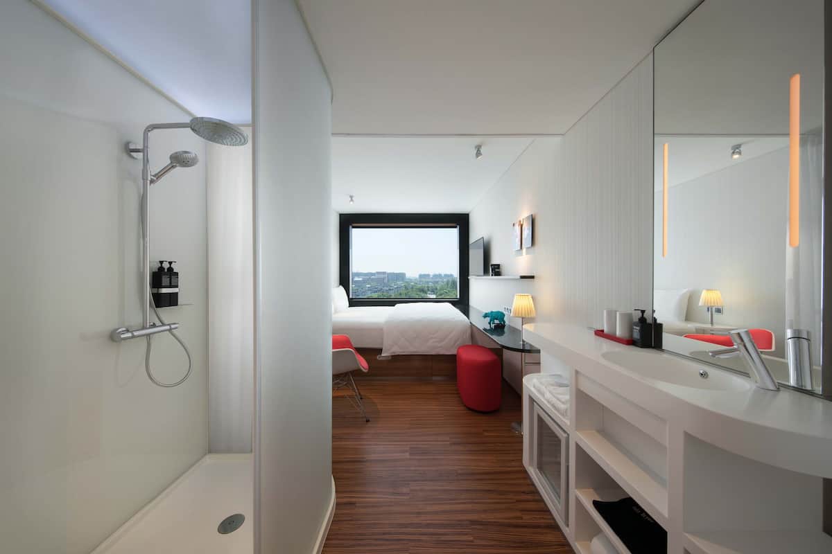 A modern hotel room with a shower, a bed, a desk, and a large window offering a city view.