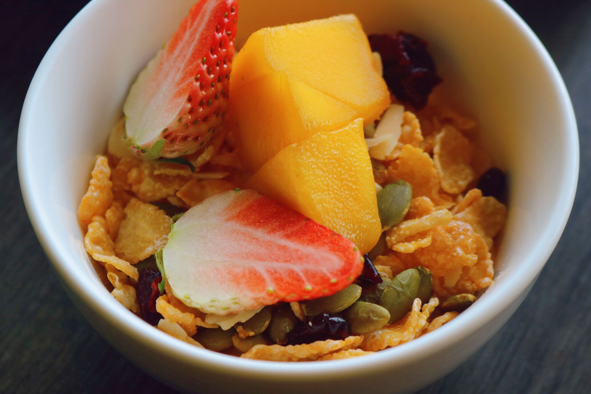 A bowl containing cereal, strawberries, mango, and pumpkin seeds.