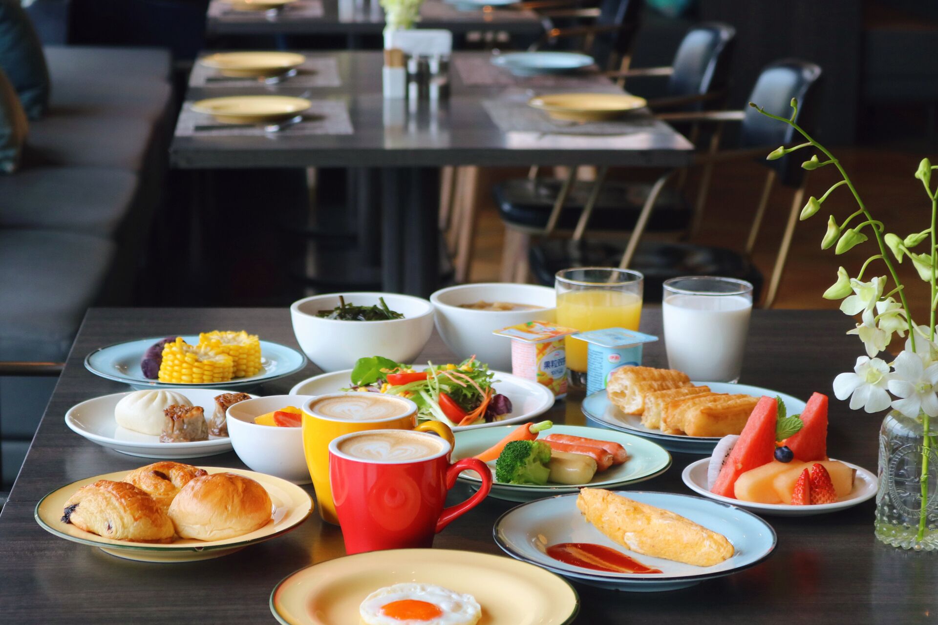 Une table à manger avec divers aliments pour le petit-déjeuner, notamment des assiettes remplies, une tasse rouge et un verre de jus d’orange.