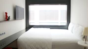 A hotel room with a large bed, a flat-screen TV mounted on the wall, a window with blinds, and a small round table with a lamp.