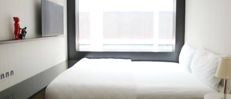 Une chambre d’hôtel avec un grand lit, un téléviseur à écran plat fixé au mur, une fenêtre avec des stores et une petite table ronde avec une lampe.