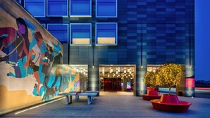 Een moderne hotelentree met een grote muurschildering, een goed verlichte gevel en terrasplekken buiten.