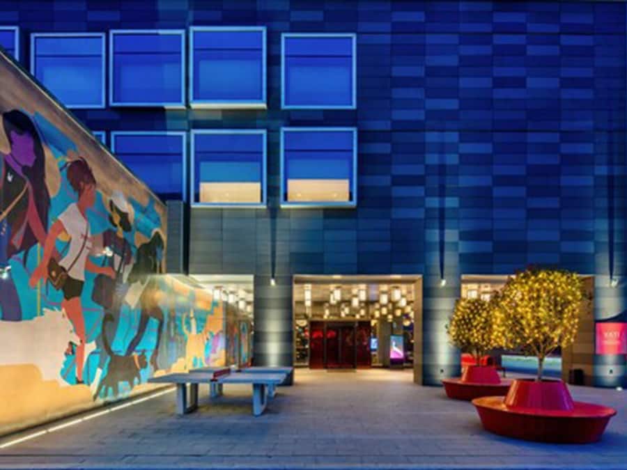 A modern hotel entrance with a large mural, a well-lit facade, and outdoor seating.