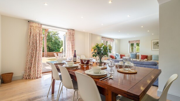 Comedor con mesa de madera dispuesta para cuatro, sillas blancas y vista al jardín a través de amplios ventanales.