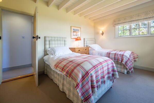 A bedroom with two beds, a nightstand, a lamp, a framed picture on the wall, and a window with a view of trees. - Alexander House, Thorpeness (Leiston)