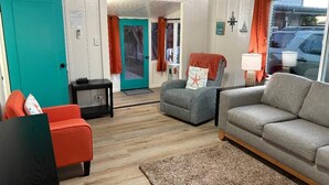 A cozy living room with a teal door, a grey sofa, an orange armchair, and a small table with a lamp. - Walk to Embarcadero + Kids Dream Yard + Pool Table (Morro Bay)