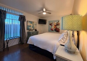 A hotel room with a bed, a ceiling fan, a TV, a painting of a cow, a lamp, and a window with curtains. - 2BR Pet-Friendly Condo, Walkable Location (Phoenix)