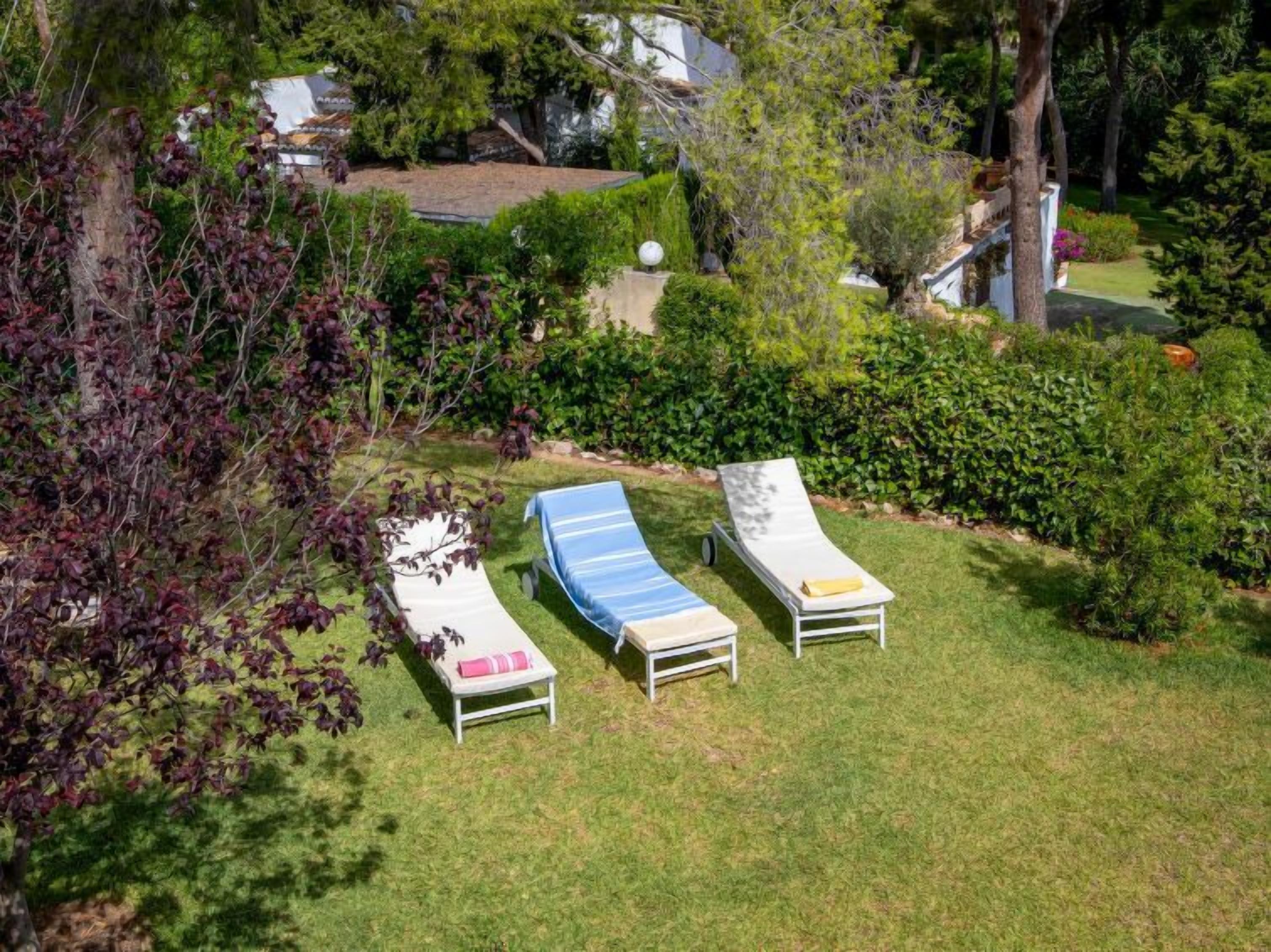 A garden with three sun loungers and a small table, surrounded by lush greenery and trees.