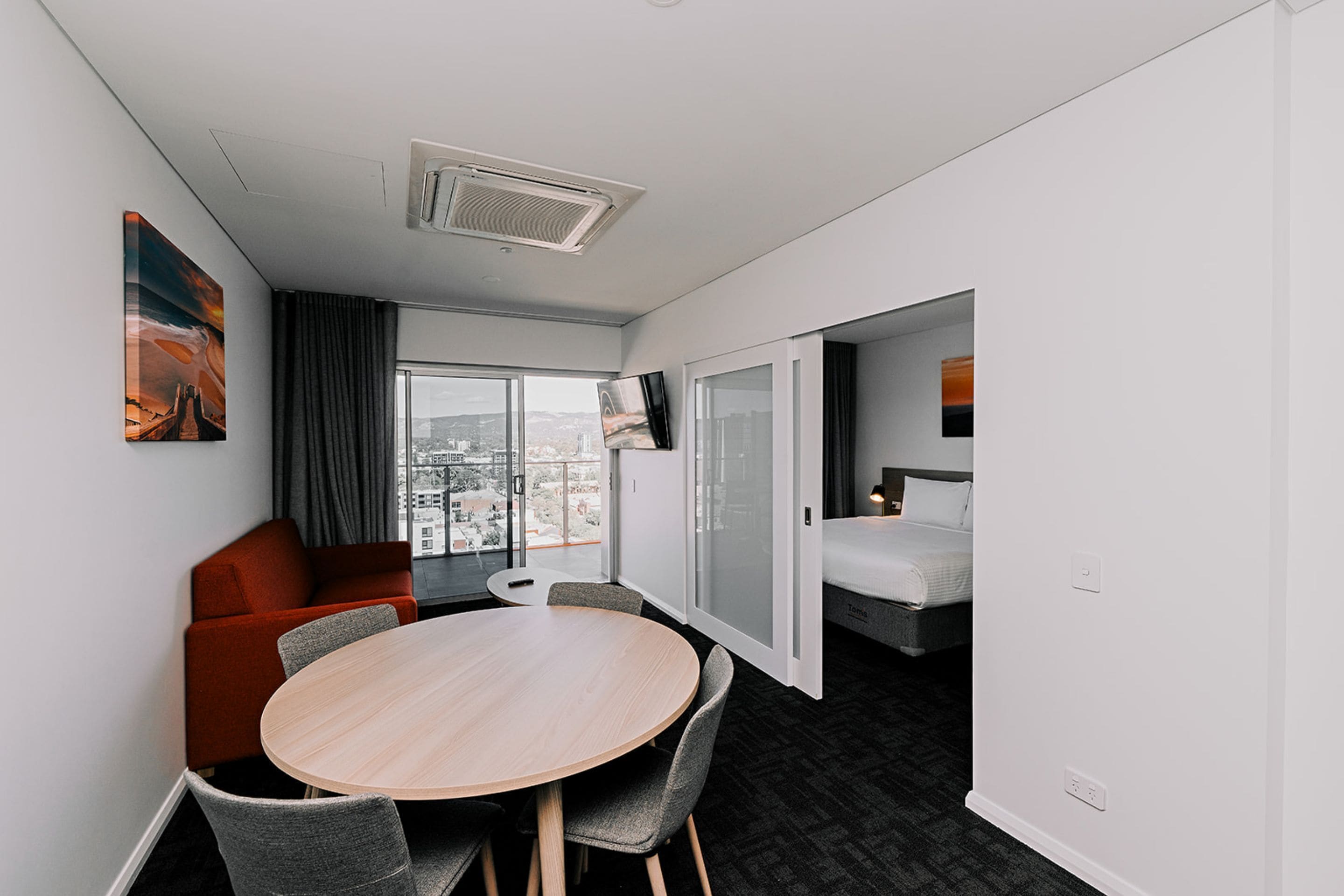 Una habitación de hotel moderna con una mesa redonda, sillas grises, un sofá rojo, una cama y un ventanal con vistas a la ciudad.