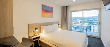 A hotel room with a bed, a desk with a laptop, a chair, a nightstand with a lamp, and a large window with a city view.