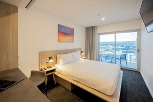 Une chambre d’hôtel avec un lit, un bureau avec un ordinateur portable, une chaise, une table de chevet avec une lampe et une grande fenêtre donnant sur la ville.