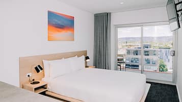 A modern hotel room with a large bed, a desk, a nightstand with a lamp, a painting of a sunset, and a view of buildings through a large window.