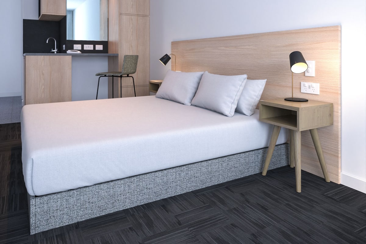 A modern hotel room with a large bed, a bedside table with a lamp, a chair, and a built-in kitchen area.