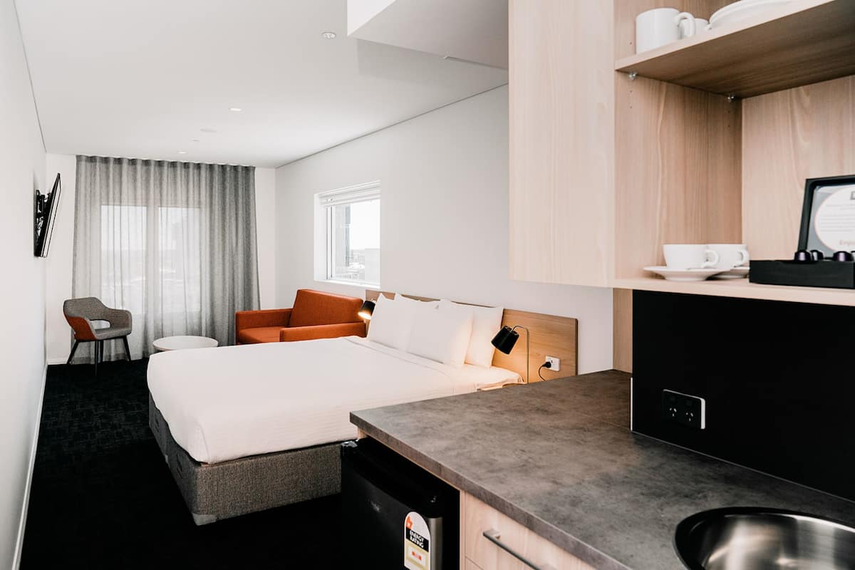 A modern hotel room with a bed, a desk, a chair, a small kitchen area, and a window with curtains.
