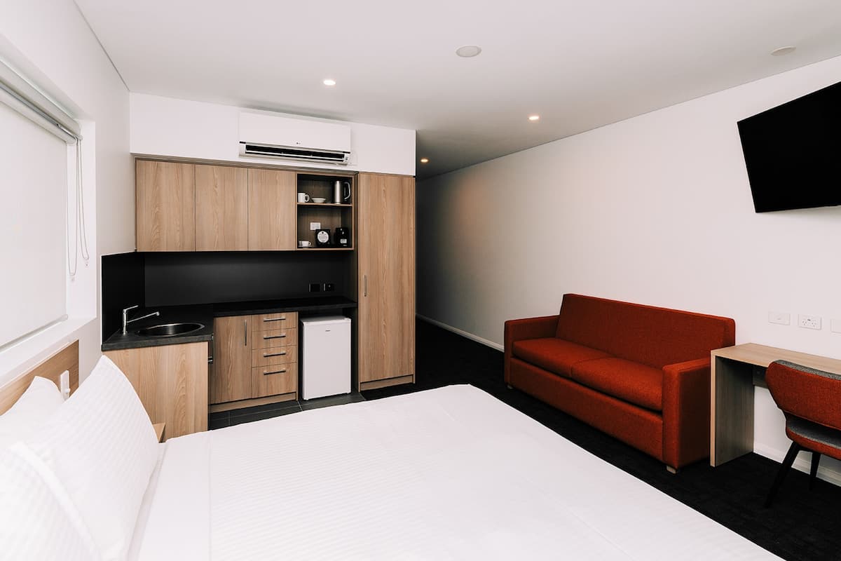 A modern hotel room with a bed, a red sofa, a small desk, and a kitchenette.