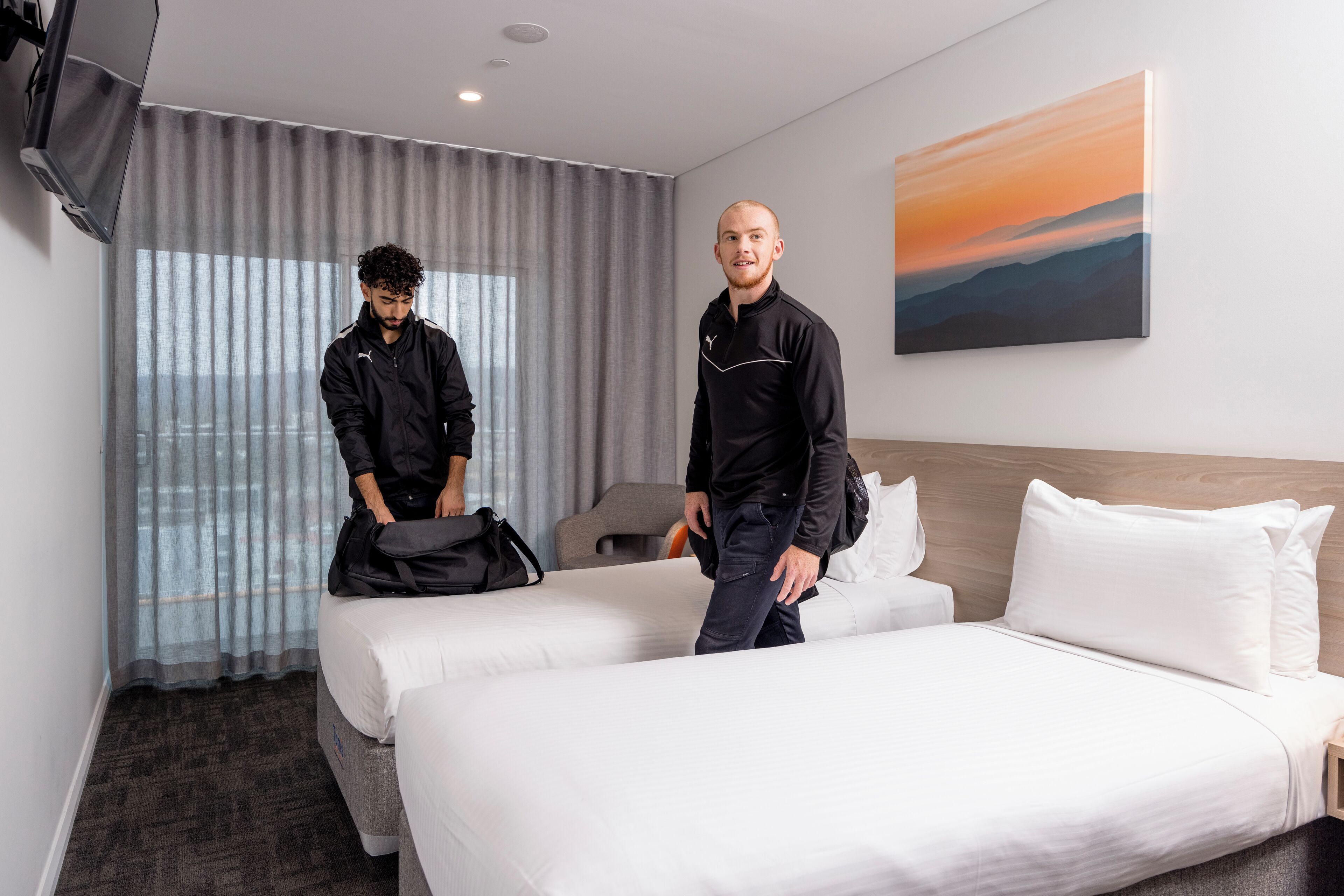 Une chambre d’hôtel avec deux lits, une grande fenêtre avec des rideaux, un tableau accroché au mur et un téléviseur fixé au mur.