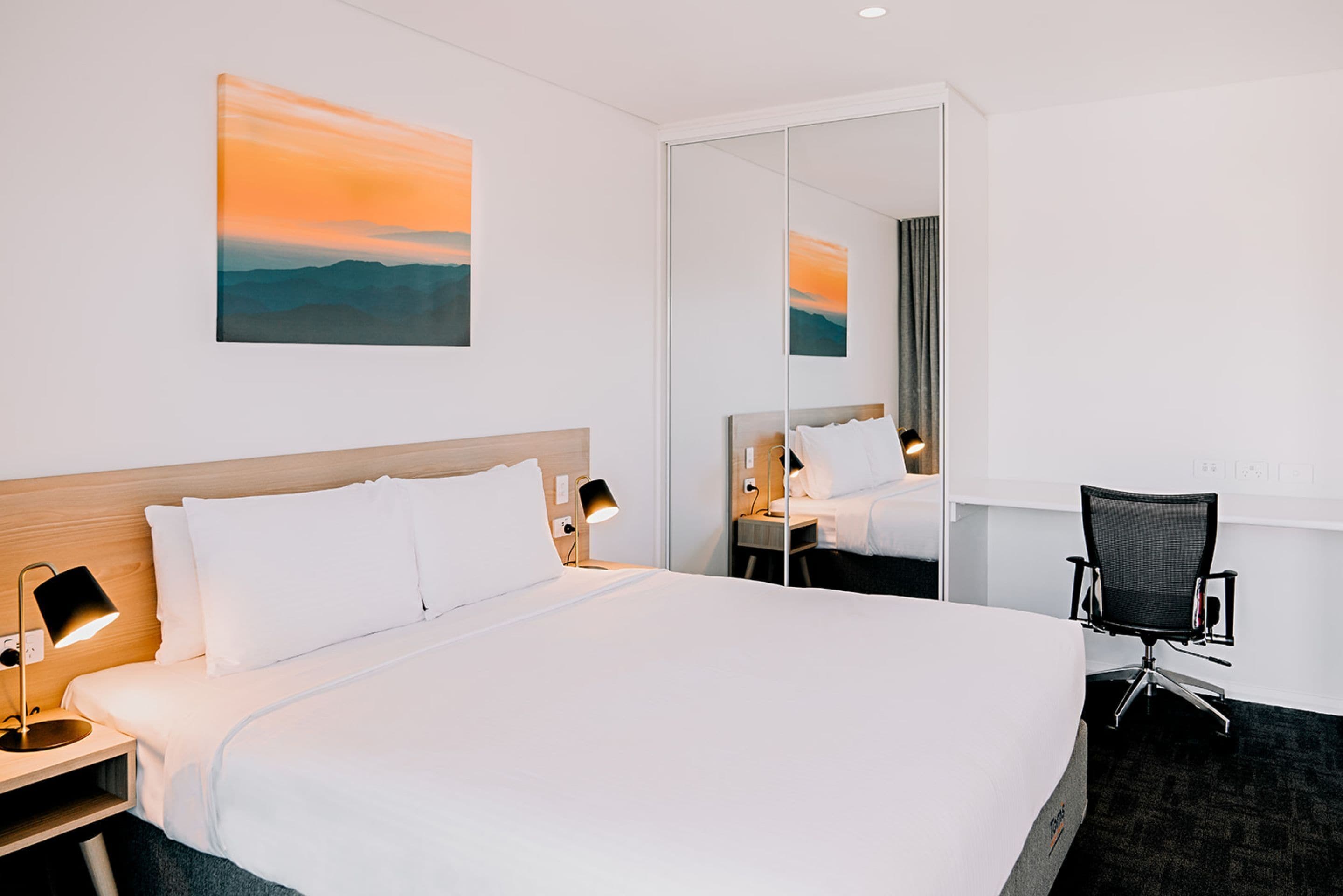 A hotel room with a large bed, a desk, a chair, a mirror, and a painting on the wall.