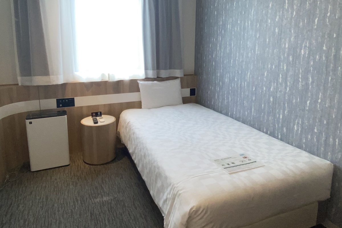 A hotel room with a bed, a small bedside table, a heater, and a window with curtains.
