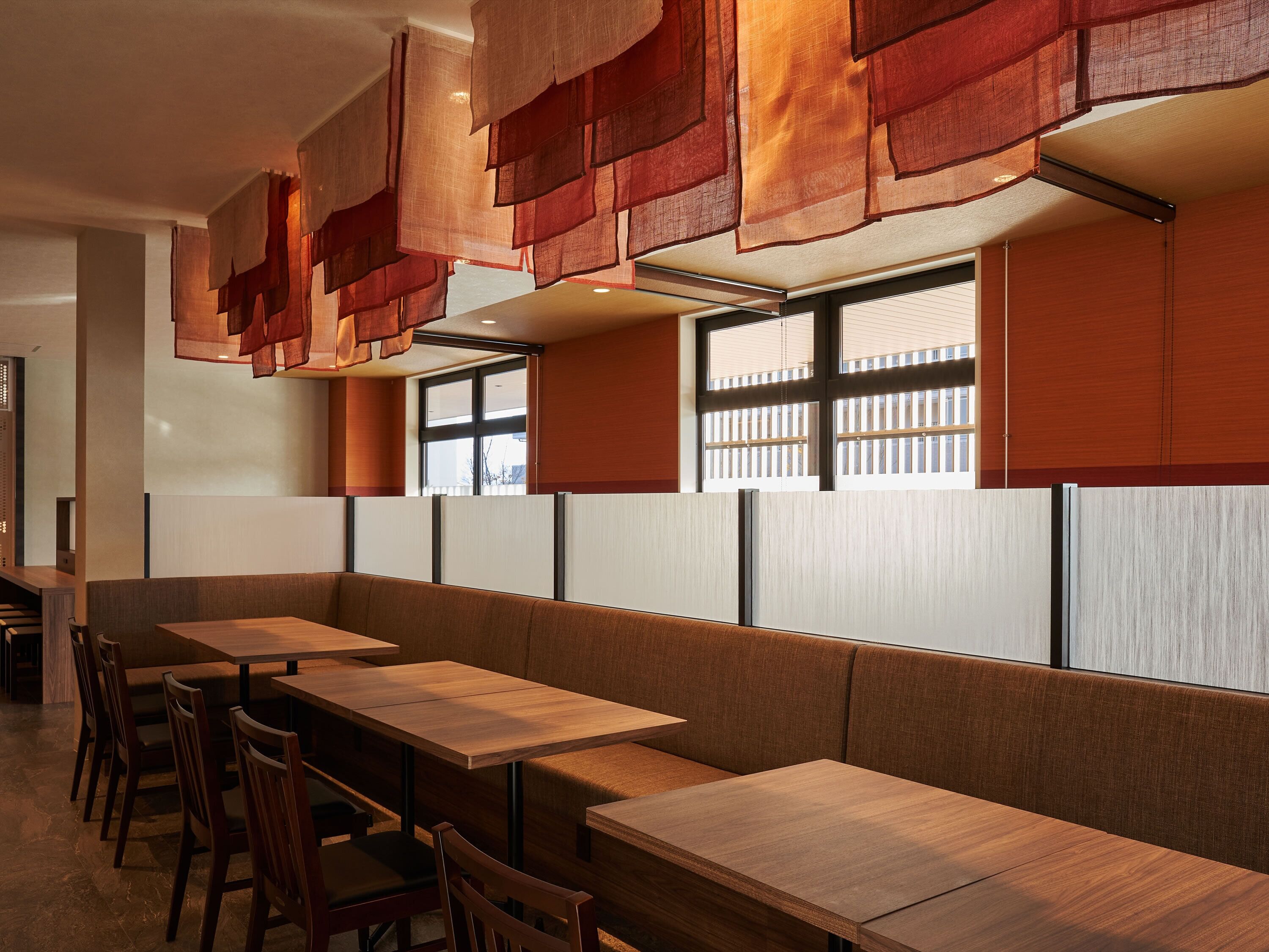 Een modern restaurantinterieur met houten tafels en banken, grote ramen met zonwering en hangende stoffen decoraties.