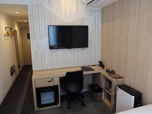 Habitación de hotel con escritorio de madera, silla de oficina negra, televisor de pantalla plana montado en la pared, mini refrigerador, cesto de basura y un calentador pequeño.