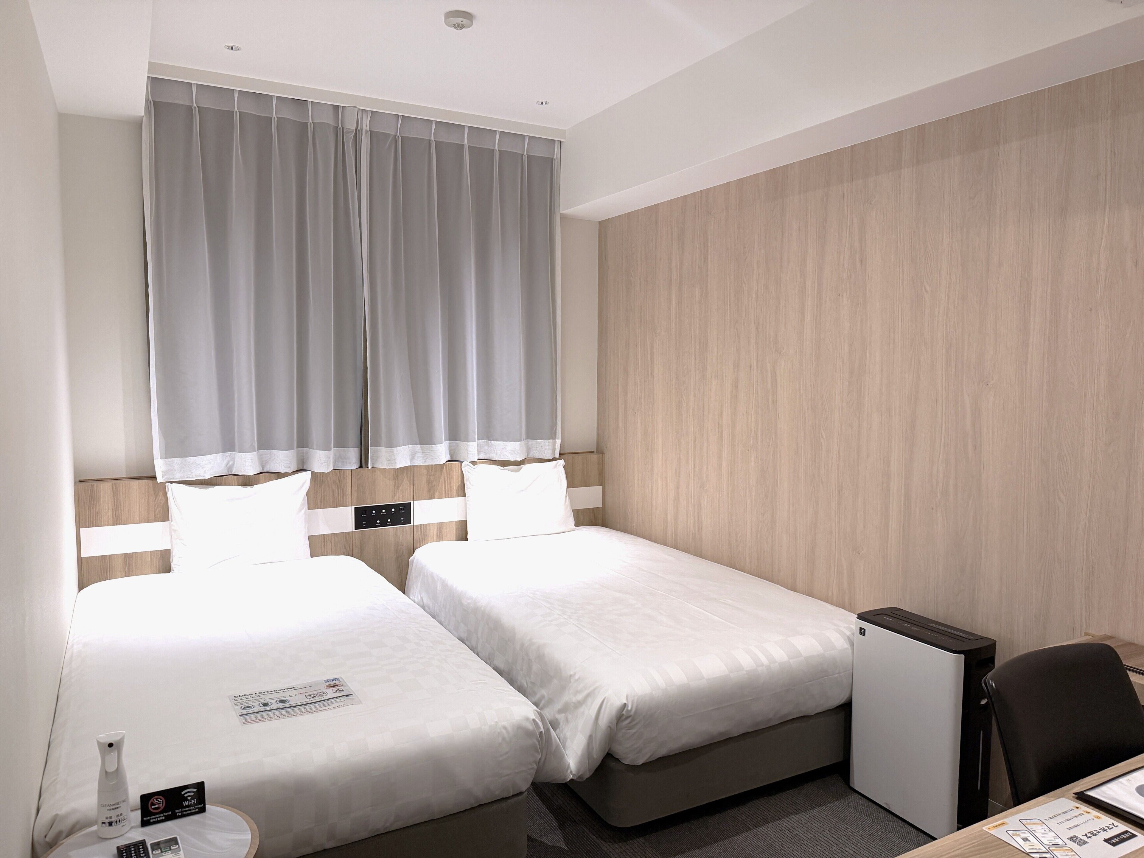 A hotel room with two beds, a desk, a chair, a small table, a lamp, and a ceiling light.