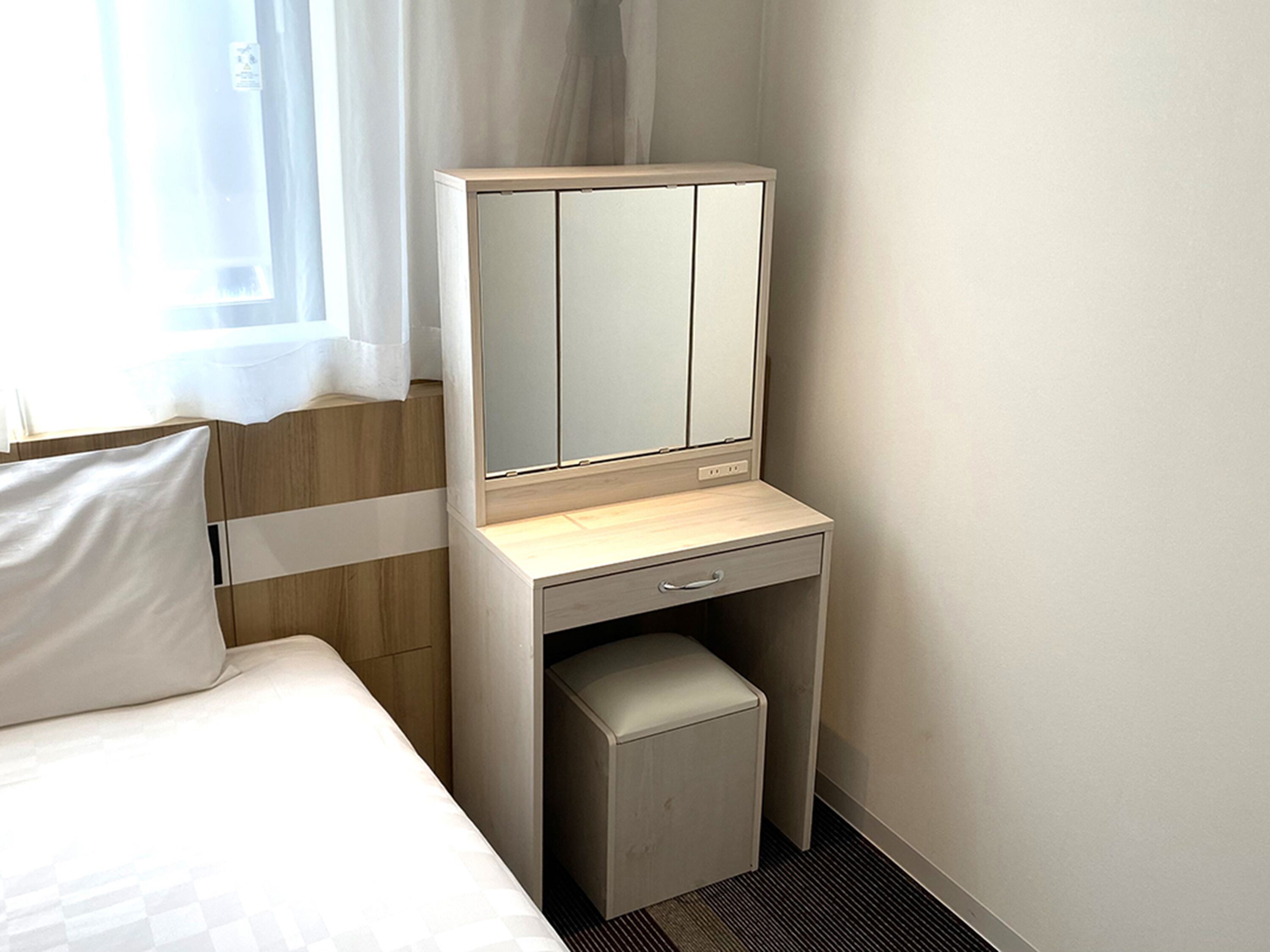 Una habitación de hotel con una cama, una mesita de noche con espejo y un pequeño cajón.
