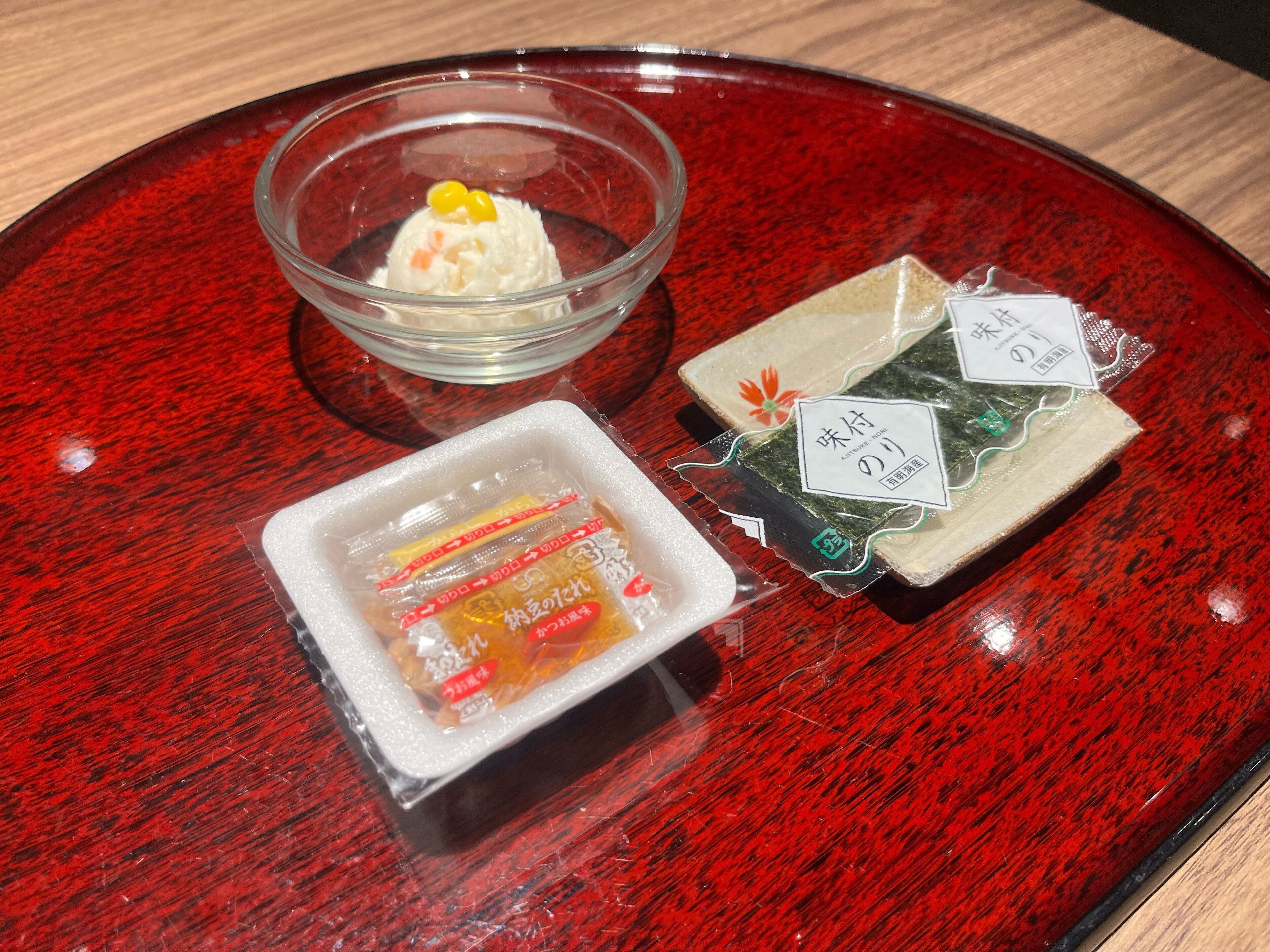 A red tray with a glass bowl containing a white dessert, two packaged snacks, and a small dish with a yellow topping.