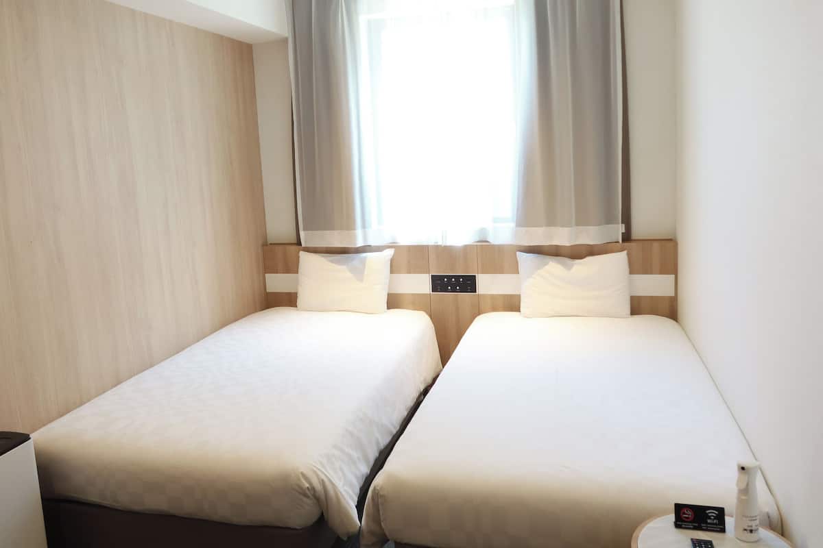 A hotel room with two beds, a window with sheer curtains, a control panel on the headboard, and a small table with a Wi-Fi sign and a spray bottle.