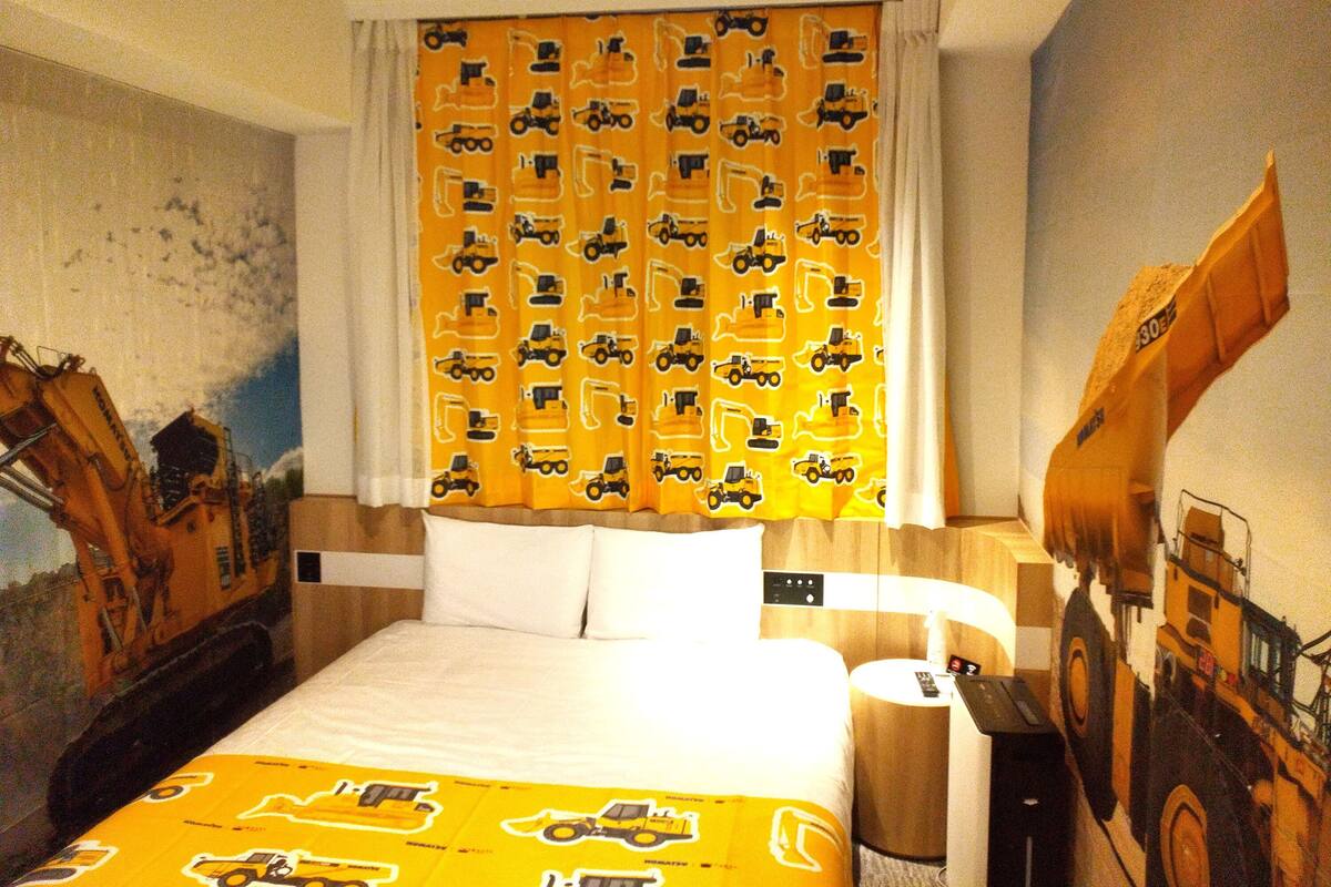 A hotel room with a bed, a nightstand, and a curtain with a construction vehicle pattern.