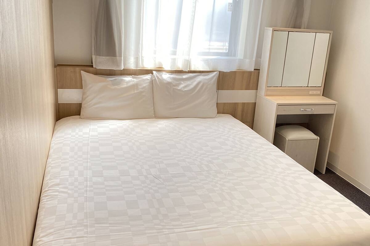 A neatly made bed with a checkered duvet, a bedside table with a mirror, and a window with sheer curtains.
