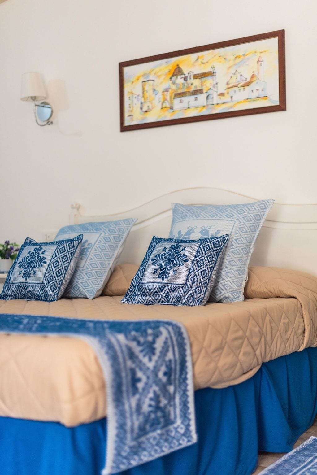 Ein Bett mit einer beige- und blauen Tagesdecke, mehrere Kissen mit blauen Mustern, eine Wandlampe und ein gerahmtes Bild über dem Bett.