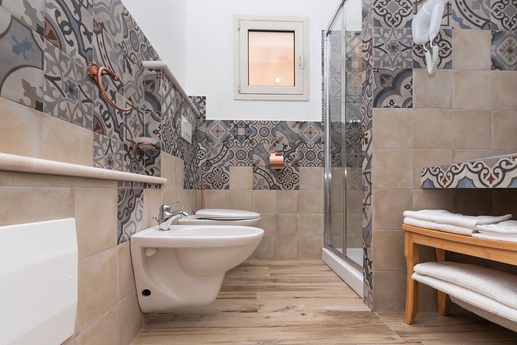 Baño con bidé, inodoro y lavamanos, con azulejos estampados y un banco de madera.