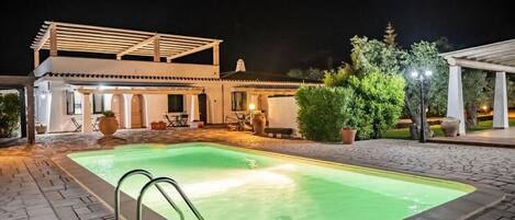 A swimming pool with a pergola, outdoor seating, and a well-lit patio area.