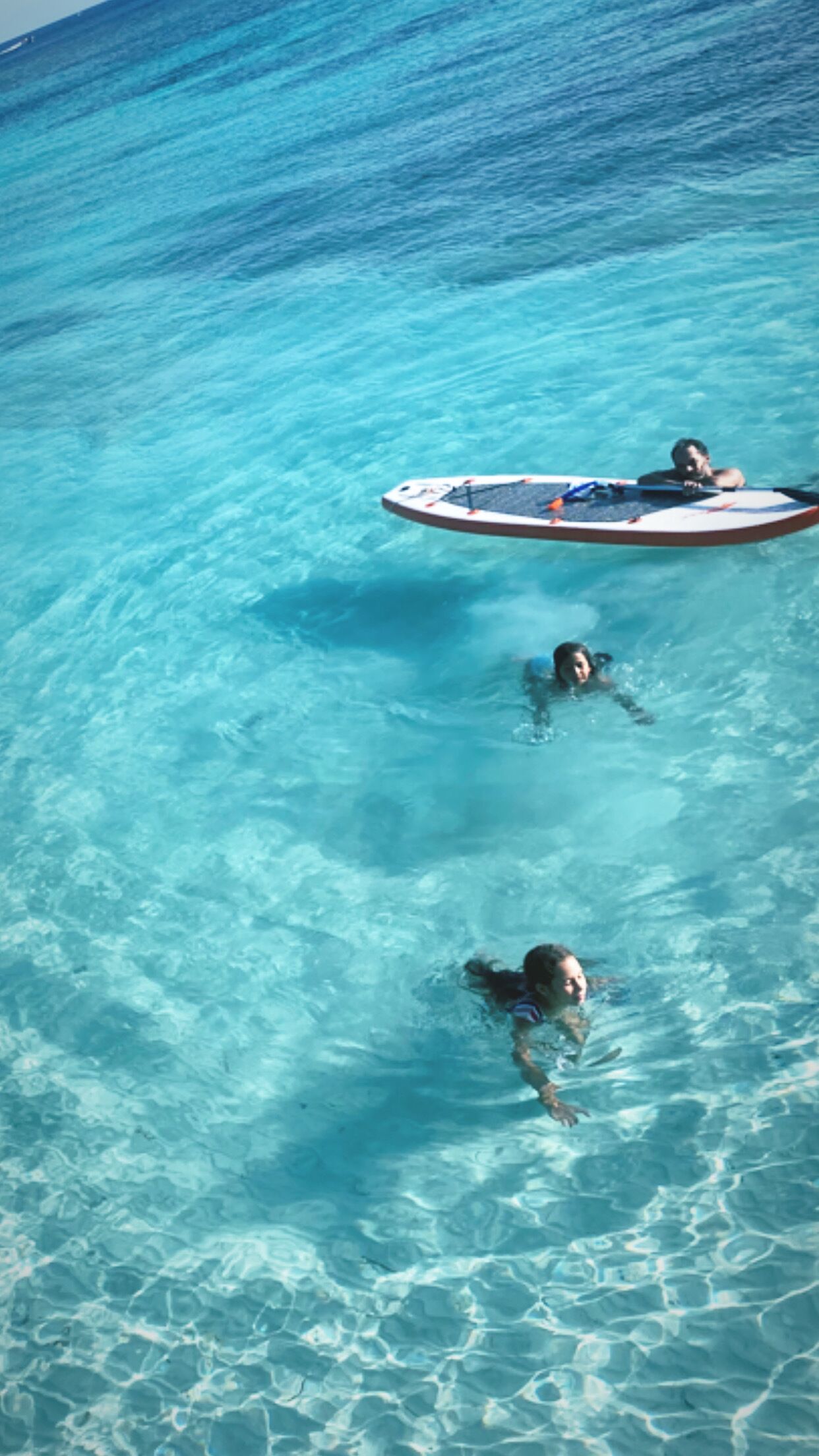 A clear blue sea with three people swimming and one person on a paddleboard.
