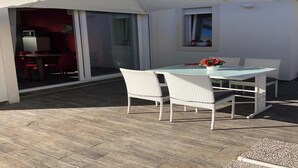 Eine Terrassengruppe im Freien mit Tisch, Stühlen, einem weißen Sonnenschirm und einer Glasschiebetür zu einem Innenbereich mit roten Sesseln.