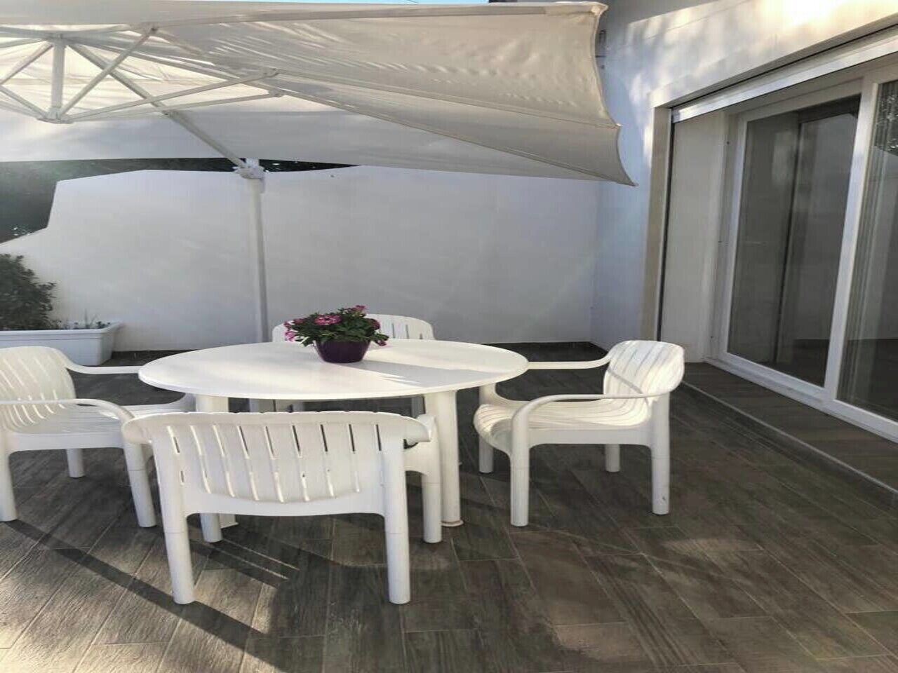 Un patio con una mesa redonda y cuatro sillas blancas, una planta en maceta y un paraguas grande.