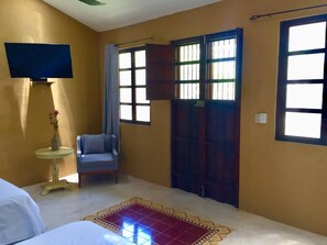 Una habitación con cama, una silla, una mesita con un jarrón, un televisor montado en la pared y una ventana con cortina.