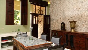 Un comedor con una mesa dispuesta para dos personas, un aparador de madera con un jarro de bronce, y una bicicleta visible a través de un ventanal abierto.