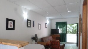 Una sala de estar con un sofá, un televisor y un ventilador. Hay una mesa con una silla y una ventana con cortinas.